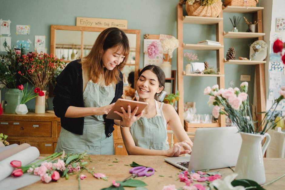 Inside a bright, cozy flower shop with soft pastel walls and wooden shelves displaying various floral arrangements and decorative items, two women are engaged in a happy conversation. One woman, stand