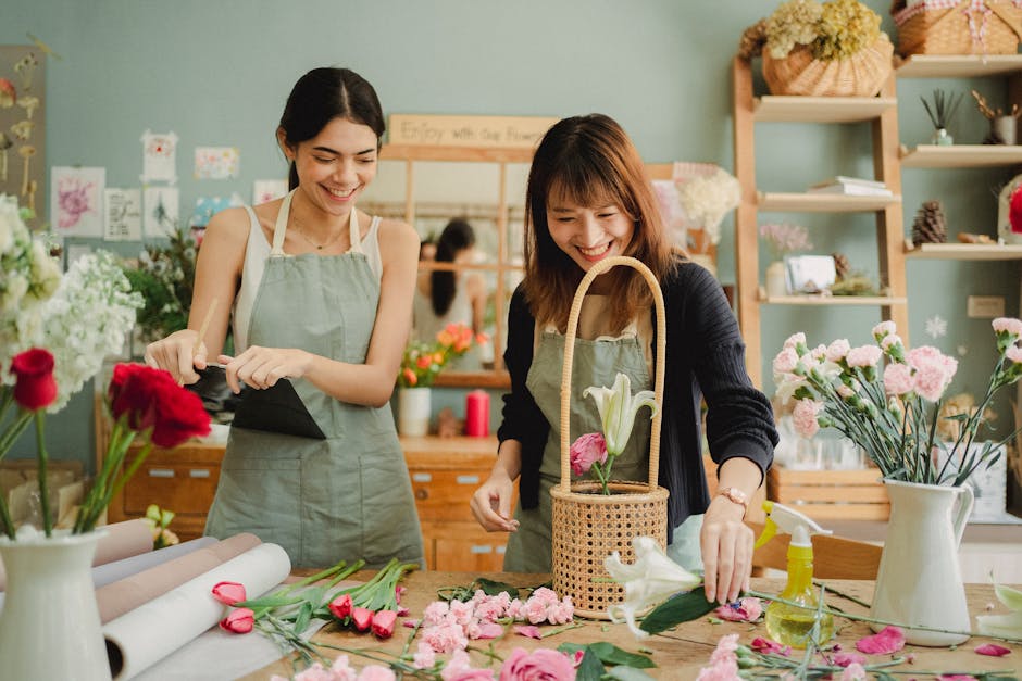 Two women working in a flower shop, arranging a floral gift with fresh white lilies and pink carnations. They are wearing aprons, smiling, and surrounded by various flowers and green foliage on a wood