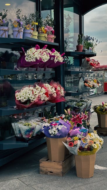 A display of fresh floral arrangements and bouquet arrangements on a multi-tiered black metal stand outside a florist shop, featuring a variety of flowers including white chrysanthemums, pink lilies, 