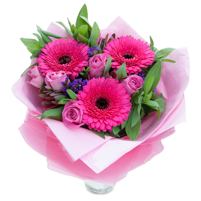 I picture this arrangement as a playful, graceful embrace in pinks and purples. At its centre three bold Gerbera daisies in a rich fuchsia burst open with confident, layered petals whose dark centres anchor the eye and lend a dramatic silhouette. Complementing them are several soft lavender-pink roses-still snug in their bud stage-whose velvety petals curl inward with delicate precision, suggesting newness and quiet romance. Interspersed among the main blooms are small sprays of purple-blue statice, which add a slightly wild, textural edge reminiscent of wildflowers peeking through a spring lawn. Surrounding all of this is a generous sweep of glossy green foliage-likely salal or eucalyptus-that provides contrast in tone and texture and brings a cool, leafy scent that balances the floral notes. The bouquet is wrapped with intention: an inner layer of pale, matte pink tissue cushions the stems while an outer veil of translucent, crinkled fuchsia-pink cellophane gives the presentation a soft, shimmering finish. The top rim of a clear glass vase shows at the base, indicating the stems have been prepared for display and that freshness is a priority. This arrangement would lift a kitchen table, brighten a bedside, or serve as a cheerful centrepiece for a small celebration in Bexley Village or near the market on the Broadway. It feels like a warm, tactile gift-soft to hold, lightly scented, and made with the exacting care of a florist who knows local streets and weekends where people like to celebrate.